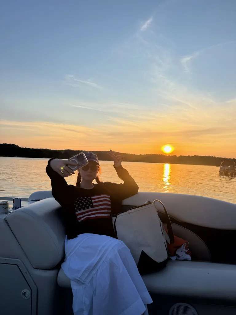 Grace Ryan on a boat wearing an American flag shirt and making a peace sign, with a sunset over a lake in the background.