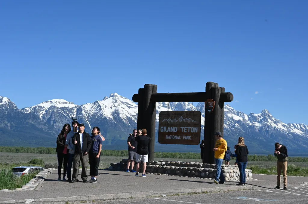 Tourists take photos at the Grand Teton National Park entrance with snow-capped mountains in the background.