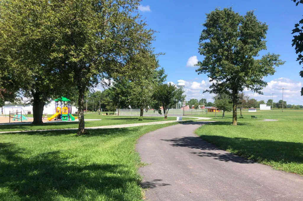 A paved trail curves through a grassy park with a playground and trees, with a water tower visible in the background.