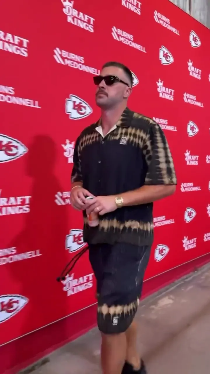 Travis Kelce wearing a tie-dye shirt and shorts, sunglasses, a watch, and a wedding band, walking in front of a red backdrop with 