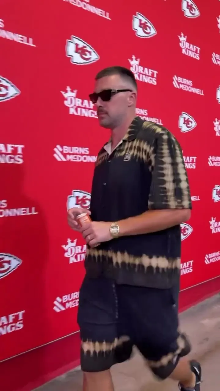 Travis Kelce walking with a drink and wearing sunglasses and a black and tan tie-dye shirt and shorts.