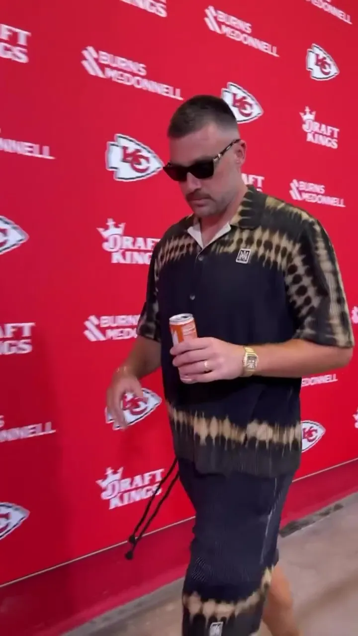 Travis Kelce wearing a tie-dye shirt, sunglasses, and a wedding band, holding a can, against a red background with team logos.