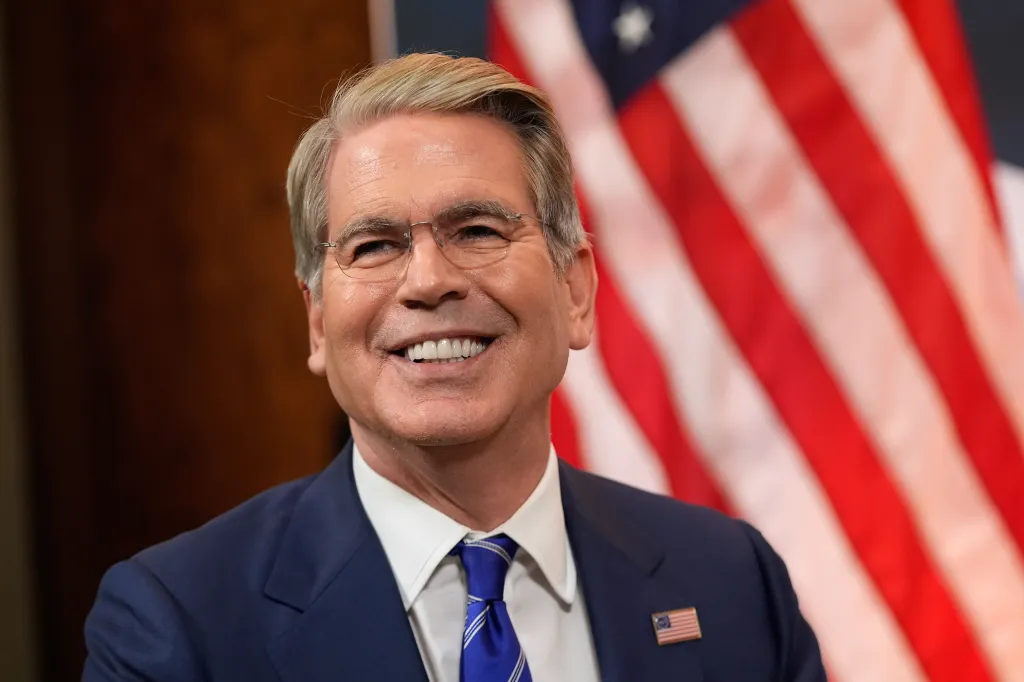 Treasury Secretary Scott Bessent smiles, with an American flag visible in the background.
