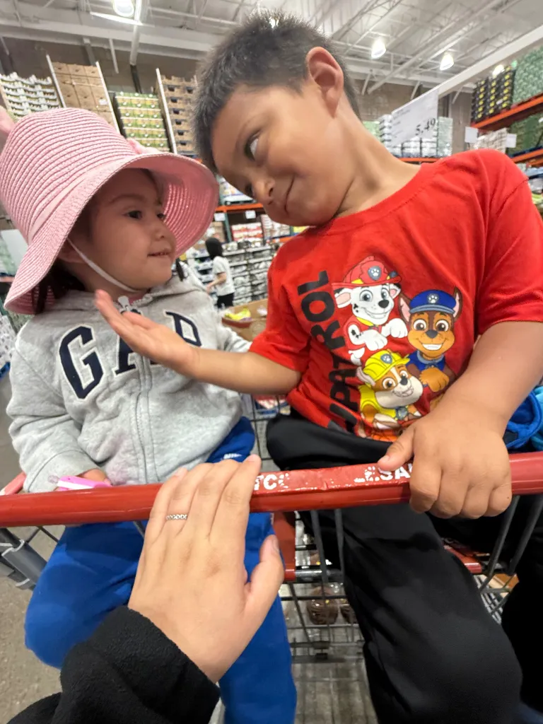 A young boy in a red Paw Patrol shirt looks at a girl in a pink hat, who is also looking at him, while both sit in a shopping cart.