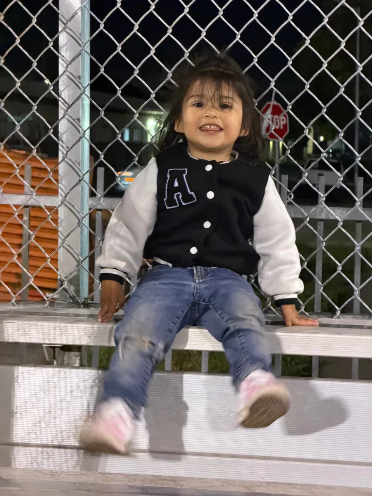 A young girl in a letterman jacket and jeans sitting on a bleacher bench.