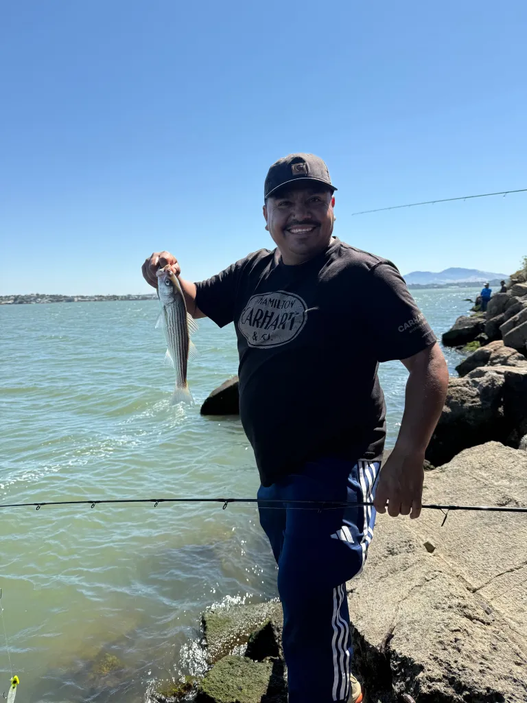 Man holding a fish by the mouth, standing on rocks next to the water, smiling at the camera.