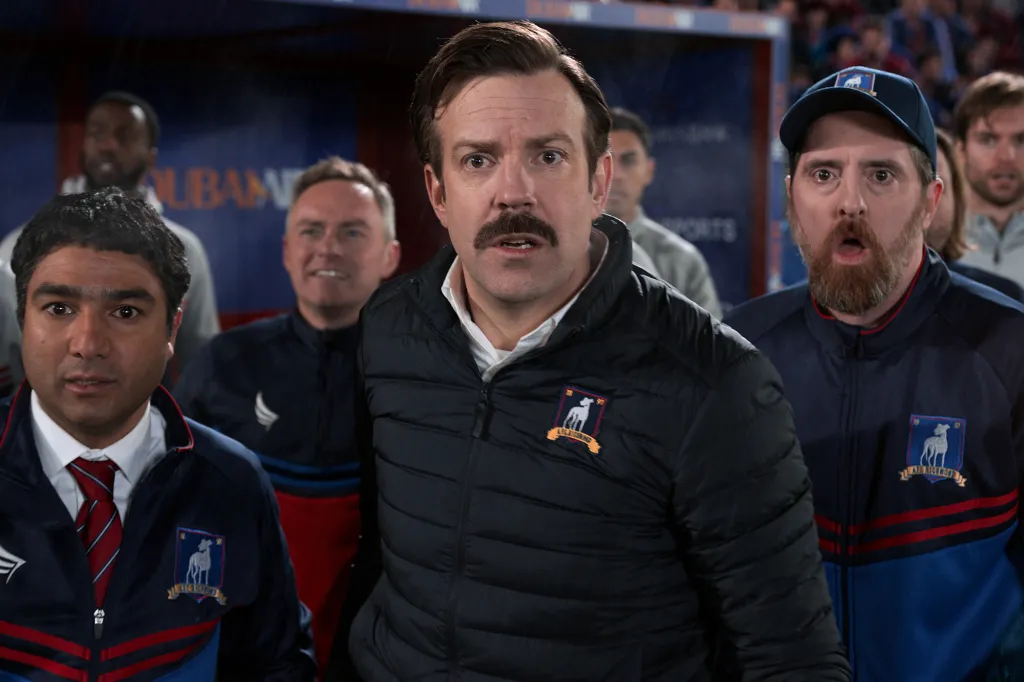 Jason Sudeikis, as Ted Lasso, with other members of the coaching staff, on the sidelines of a soccer game.