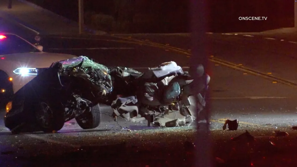 Wreckage of a car that split in half after a fatal crash, with a police car's flashing lights visible in the background.