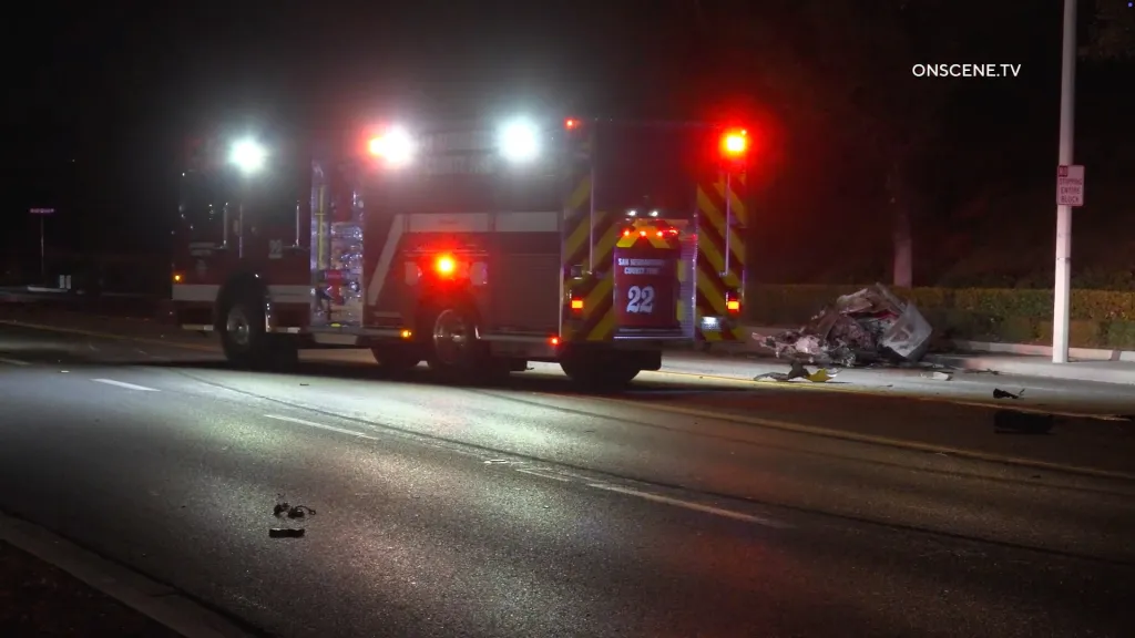 A fire truck with flashing lights parked next to the remains of a car crash on a dark road.