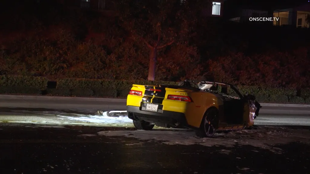 A yellow Chevrolet Camaro with black racing stripes on its trunk is shown crashed on a dark road at night.
