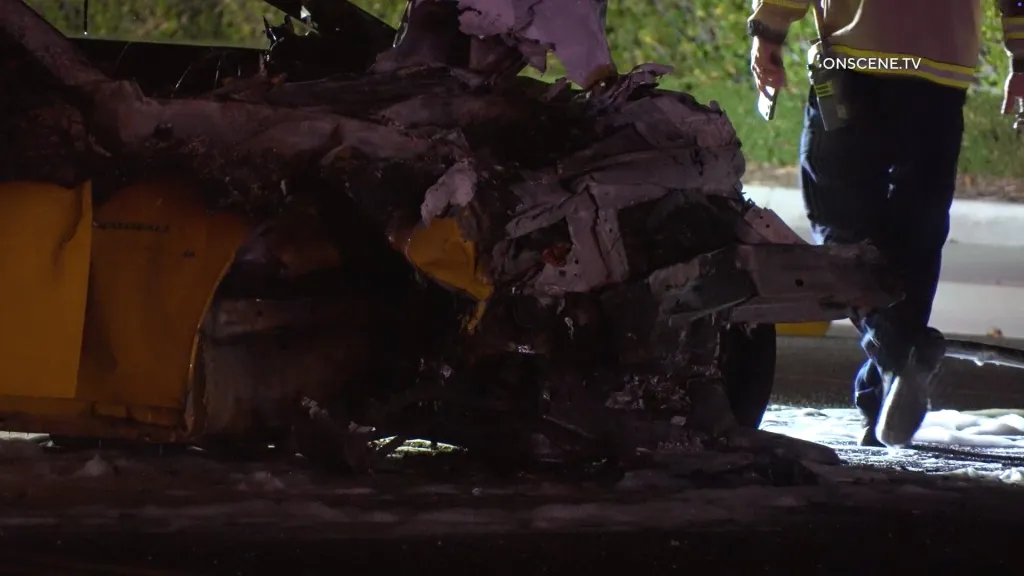 Wreckage of a yellow car, with a person in a uniform standing nearby.