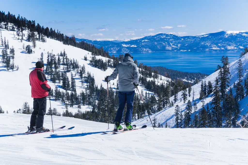 Two men on skis overlook Lake Tahoe and snow-covered mountains.