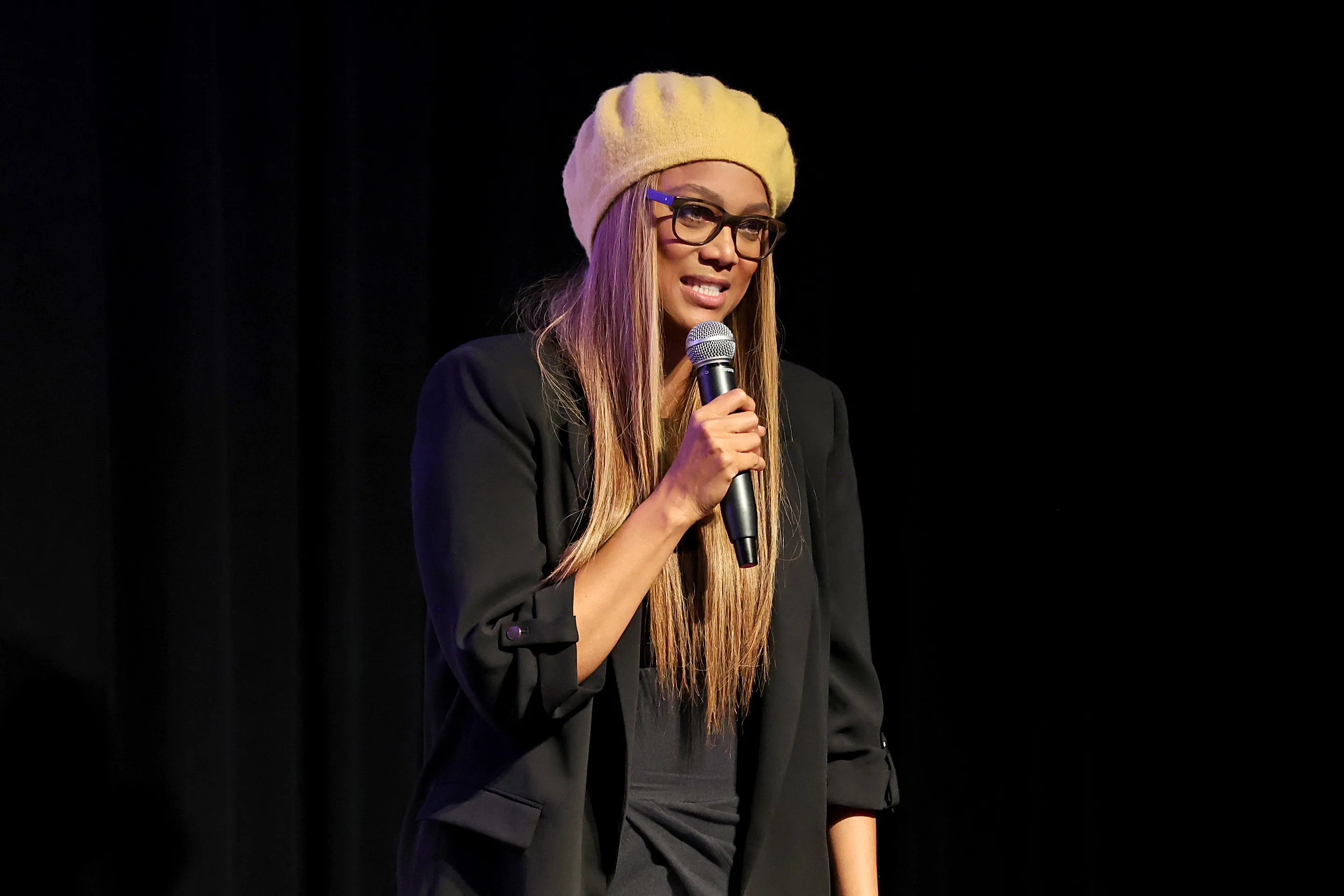 Tyra Banks speaking into a microphone while wearing a yellow beret and glasses.