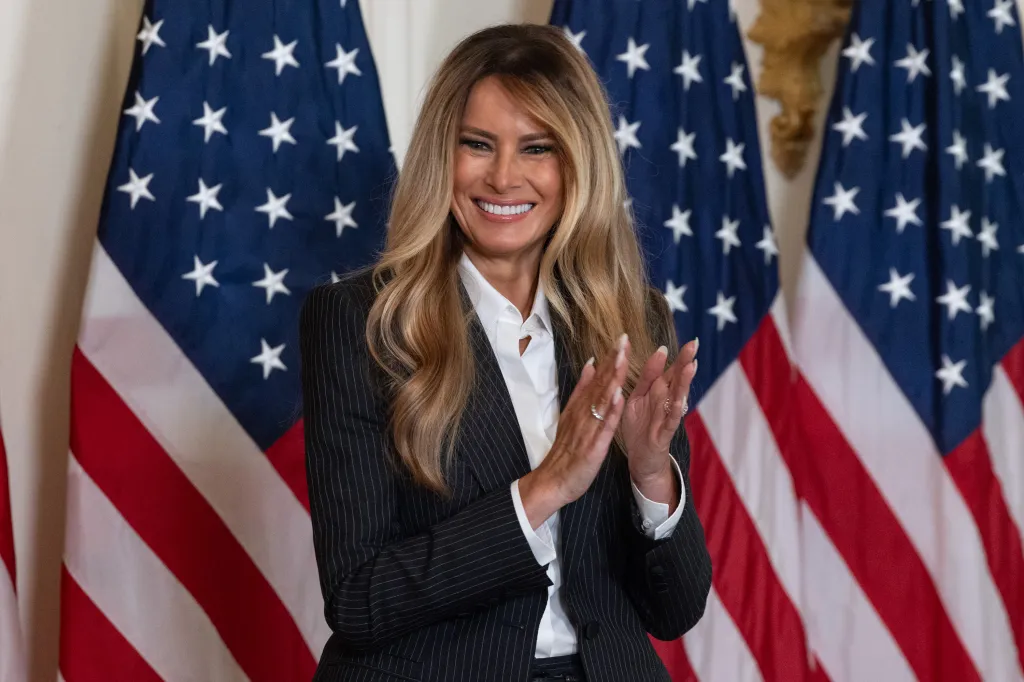 First Lady Melania Trump clapping in front of two American flags.