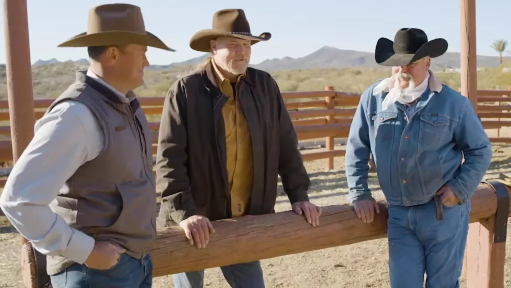 Three men in cowboy hats and western wear lean on a wooden fence outdoors.