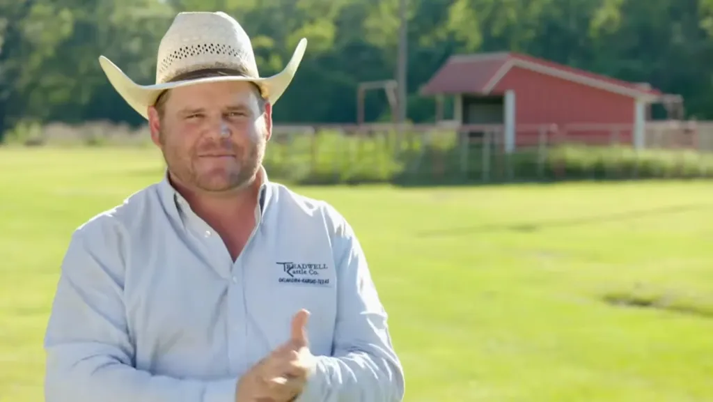 A cowboy in a hat and light blue shirt in a field with a red barn in the background.