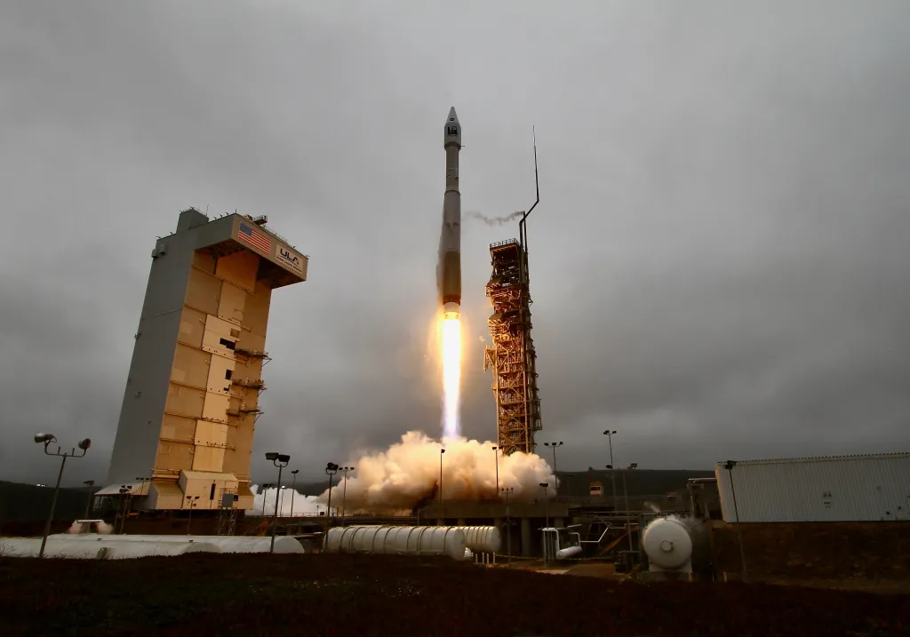 A United Launch Alliance Atlas V rocket lifting off with bright orange exhaust against a cloudy sky, next to a tall grey and tan building and a launch tower.