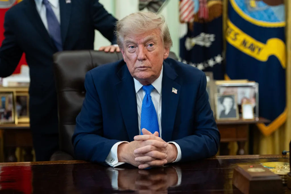 Donald Trump sits at a desk in the Oval Office, hands clasped, with another person standing behind him.