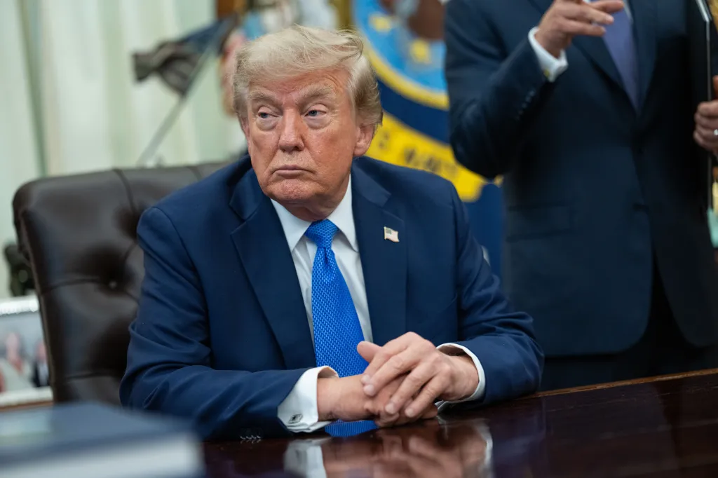 Donald Trump in a blue suit and tie, sitting with clasped hands at a desk in the Oval Office.