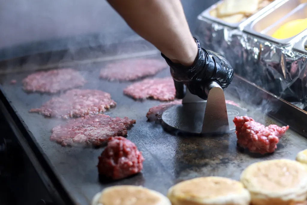 A person in black gloves smashes raw burger patties on a hot grill.