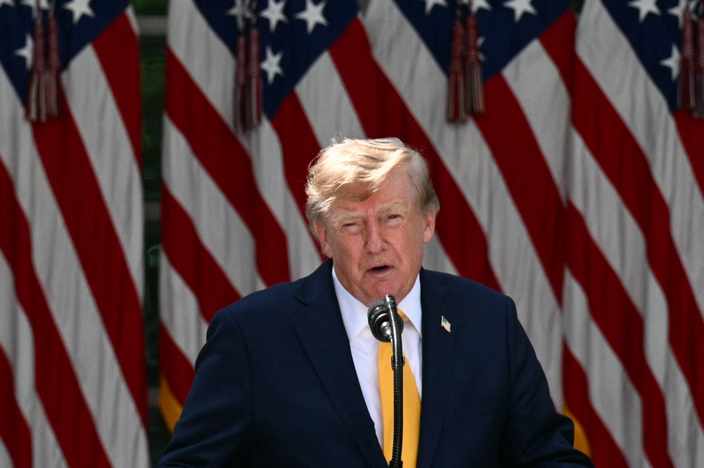 US President Donald Trump speaking at a microphone with American flags in the background.