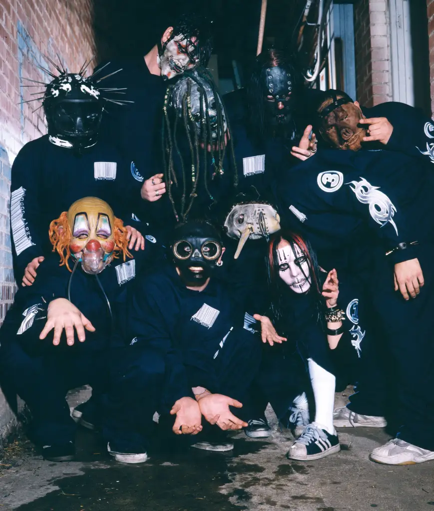 A group of Slipknot band members wearing masks and dark jumpsuits, posing against a brick wall.