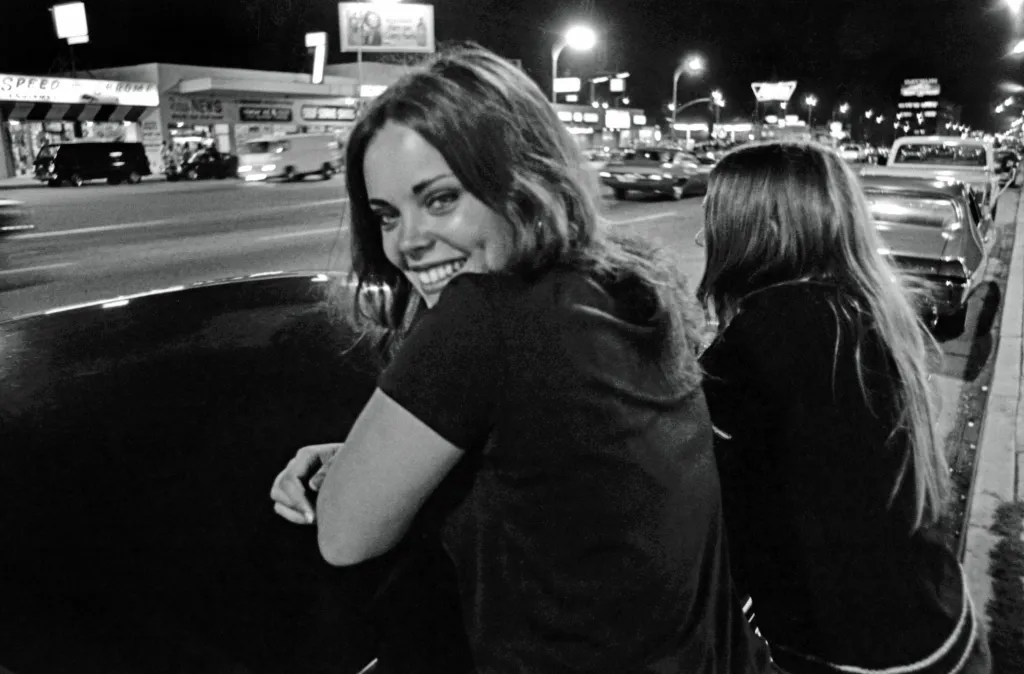 Black and white photo of a woman smiling at the camera, with another woman and a street scene with cars and businesses in the background, at night in Van Nuys, CA.