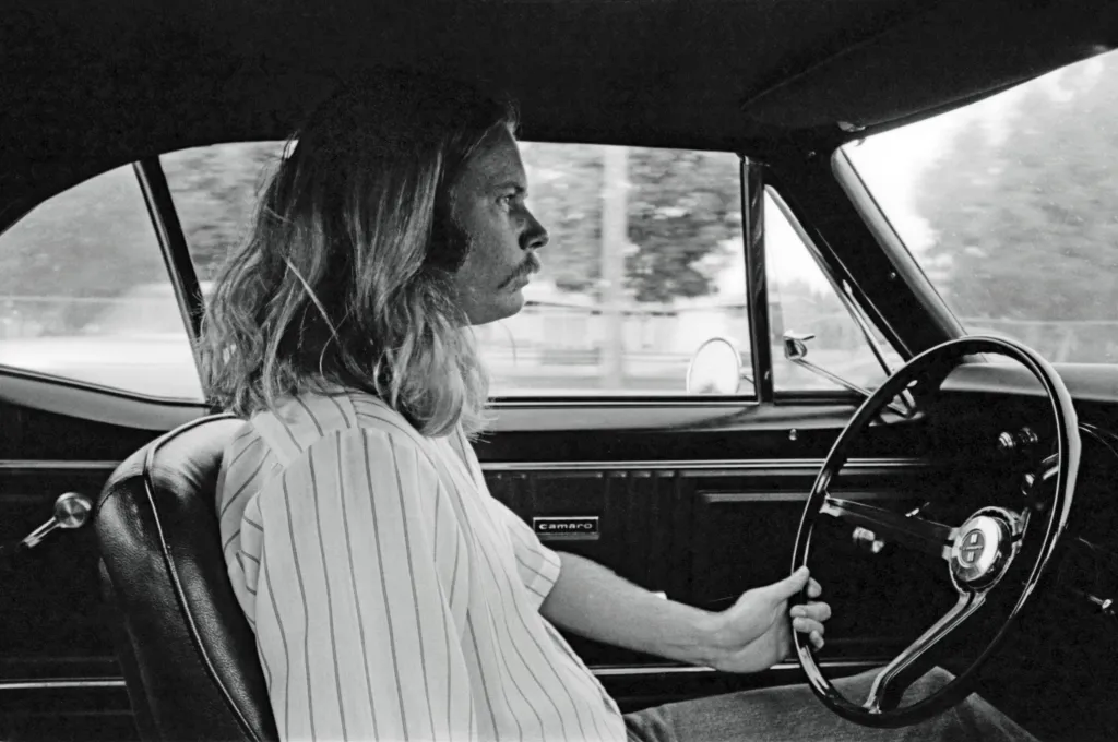 A man with long hair and a mustache driving a car in Van Nuys, CA.