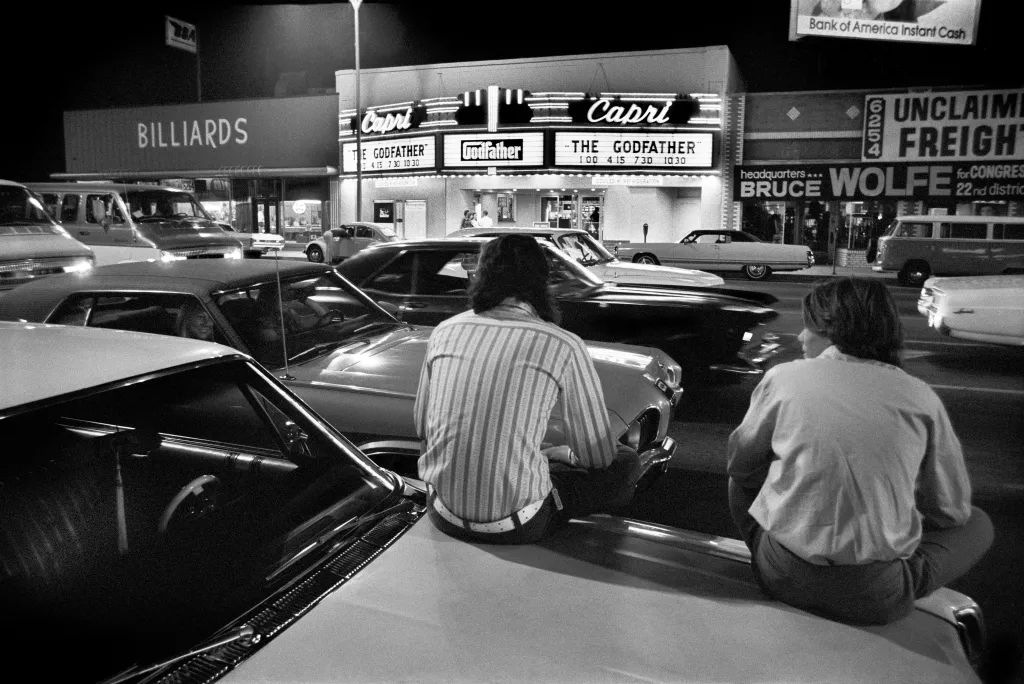Two people sit on the hood of a car with other cars and a movie theater playing