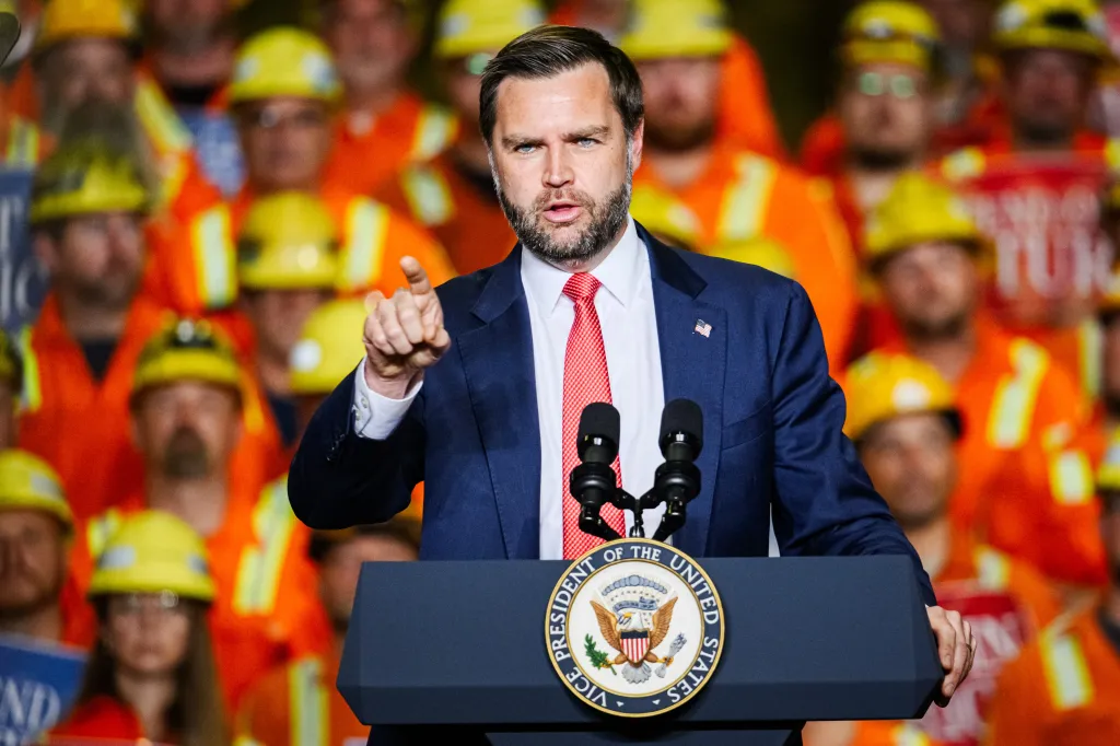 Vice President JD Vance speaking at a podium with a crowd of workers in hard hats behind him.