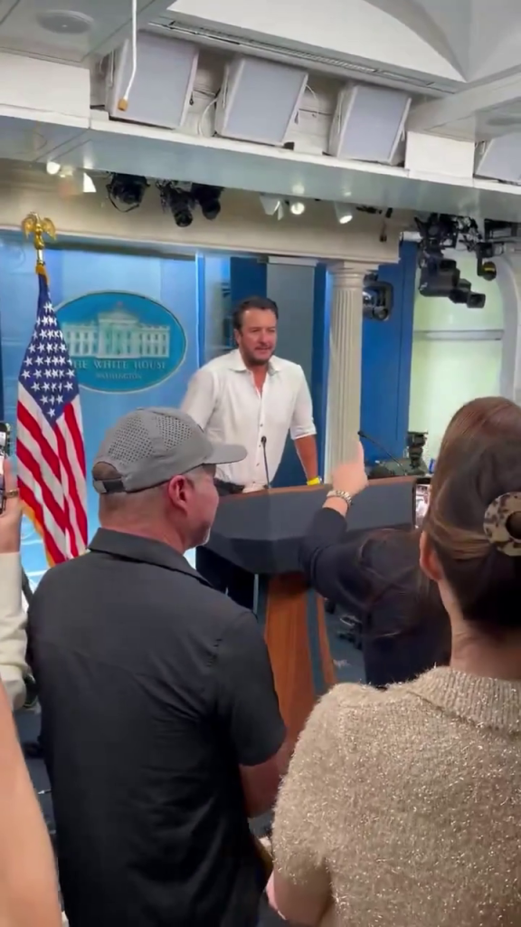 Luke Bryan at a White House press briefing podium, surrounded by people holding up phones.