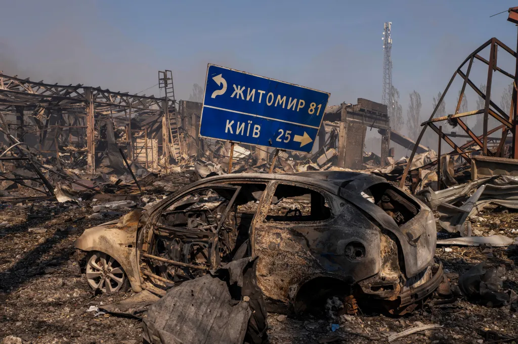 A destroyed car and damaged buildings with a road sign indicating directions to Kyiv and Zhytomyr.