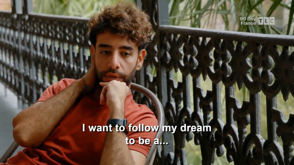 A man with curly brown hair and a beard sits on a balcony, with the caption