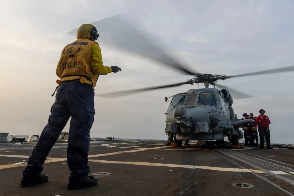 A crew member in a yellow vest and helmet stands on a warship deck facing a helicopter with rotating blades.