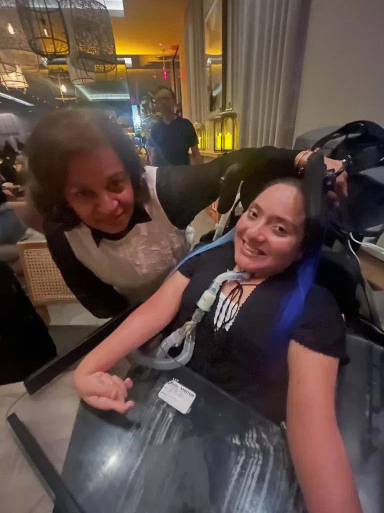 A woman with a medical device around her neck and blue hair, smiling, while another woman leans over her from behind.