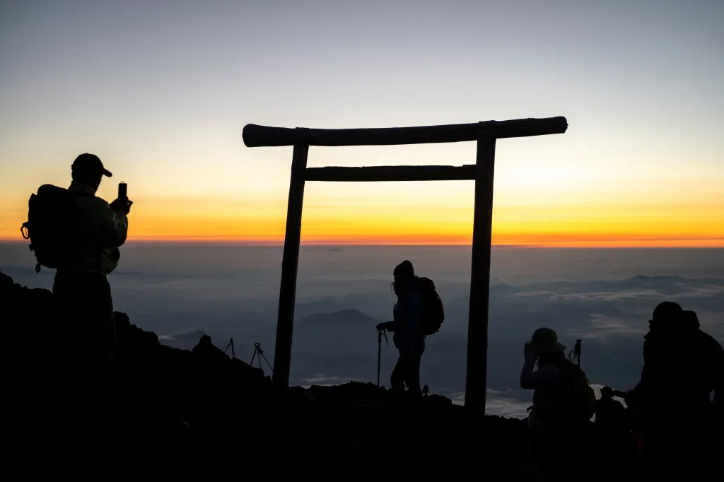 Silhouetted people watching the sunrise from the summit of Mount Fuji, with a torii gate in the middle.