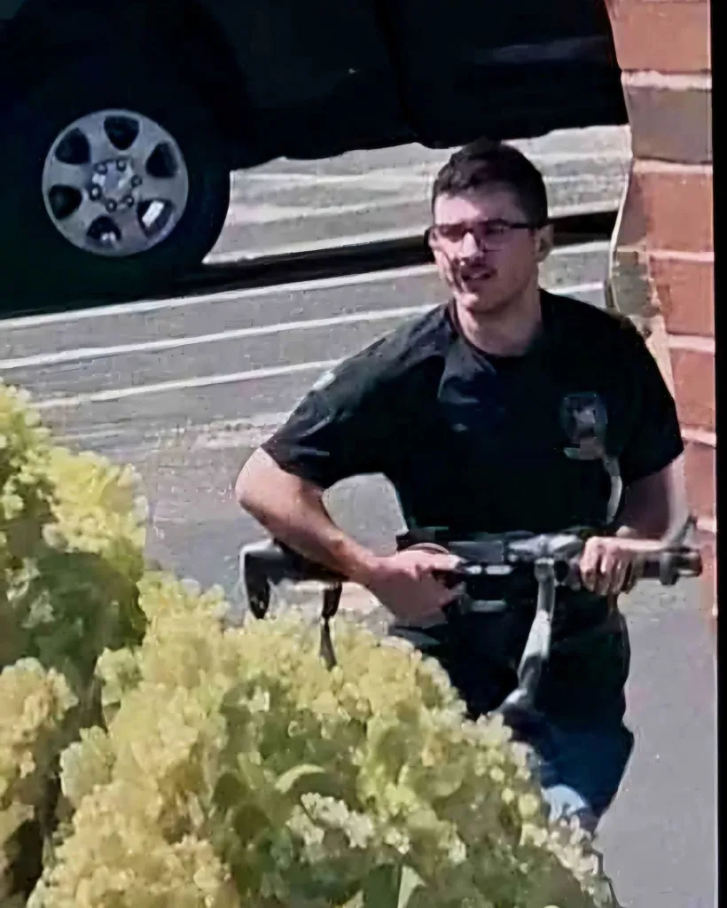 Man with a rifle and glasses, wearing a dark short-sleeved shirt, standing near a plant and a brick wall.