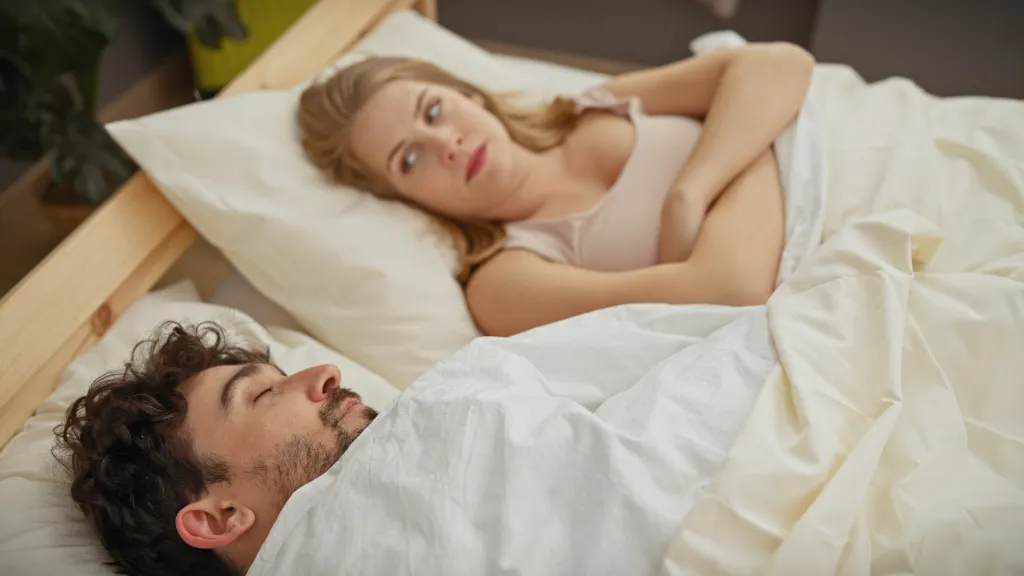 A woman lies awake in bed next to a sleeping man, depicting insomnia.