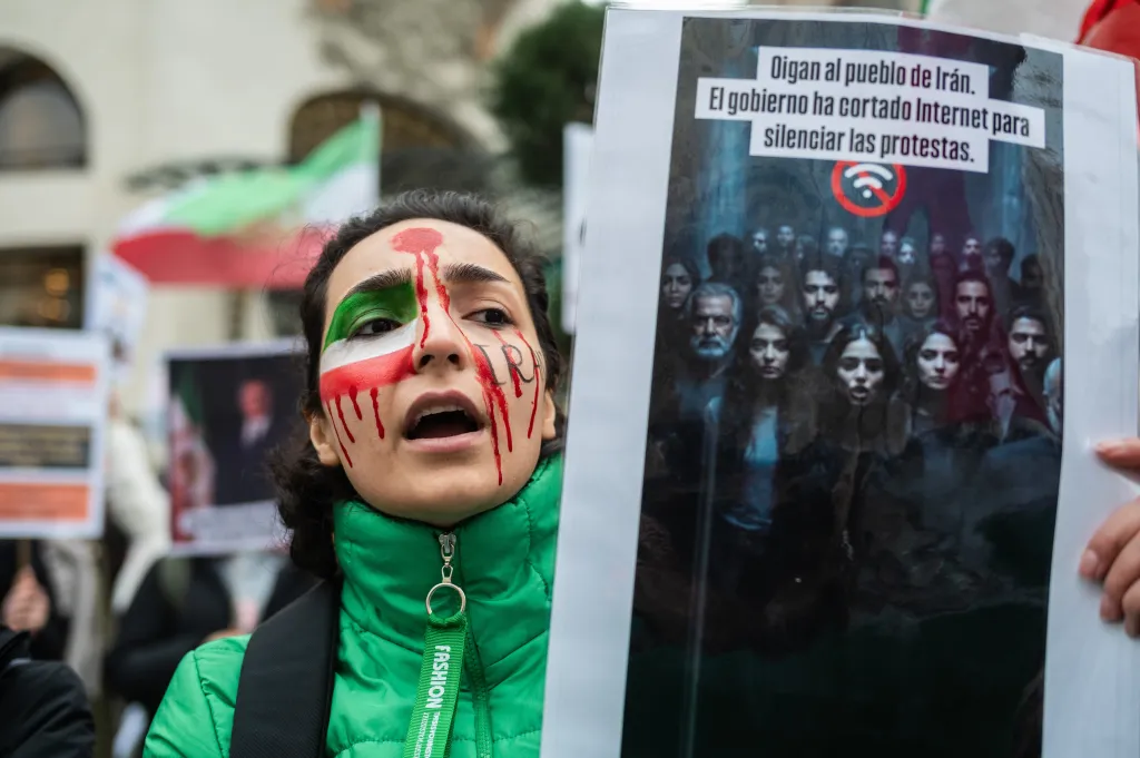 A woman with her face painted like the Iranian flag and red streaks resembling blood, holds a sign that reads, 