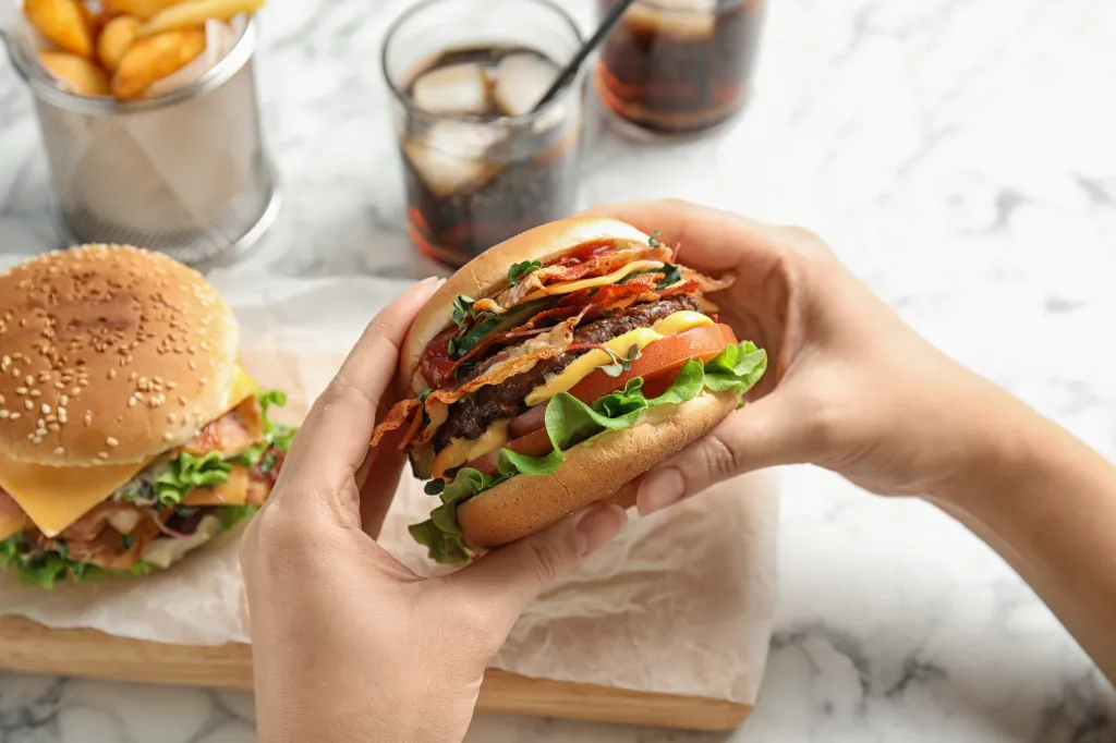 Woman holding a cheeseburger with bacon, lettuce, tomato, and cheese over a wooden board.