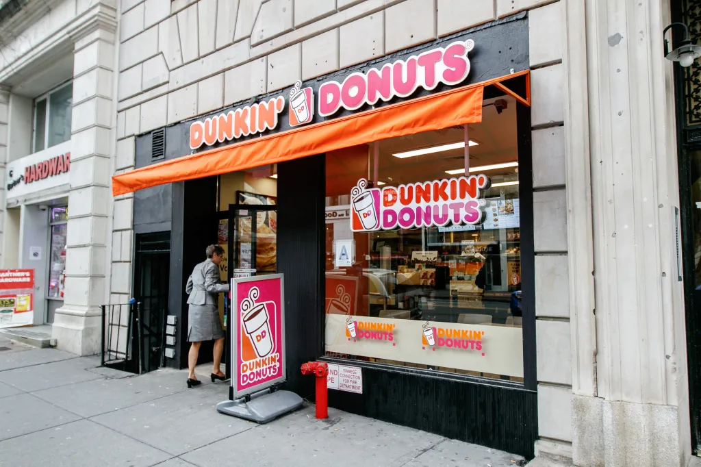 A woman walks into a Dunkin' Donuts store with an orange awning and a pink and orange sign.