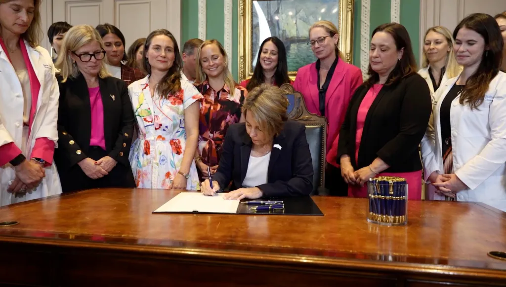 Massachusetts Governor Maura Healey signing legislation, surrounded by smiling women.