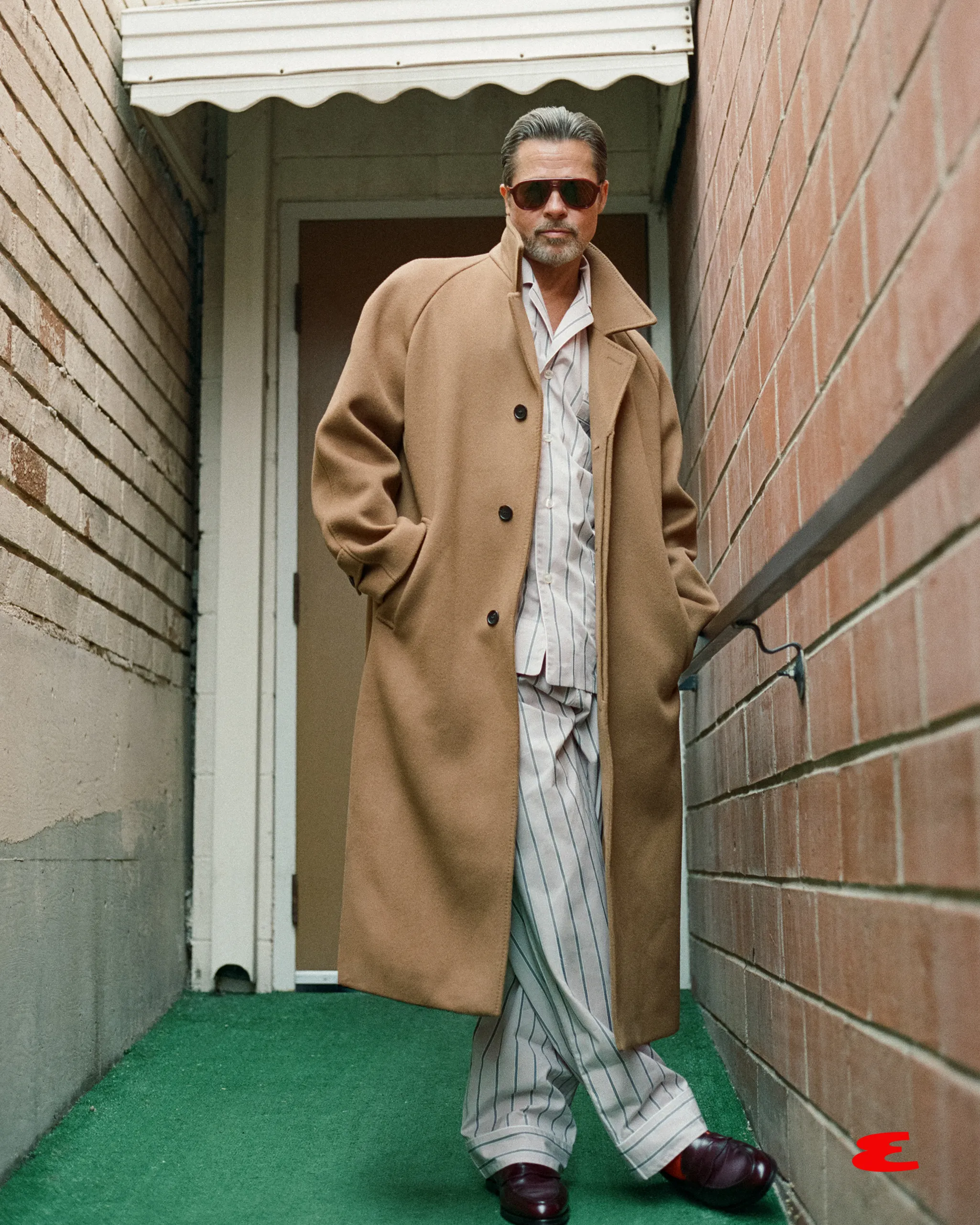 Brad Pitt wearing a long tan coat, striped pajamas, and sunglasses, leaning against a brick wall.