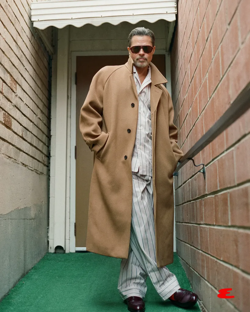 Brad Pitt in a tan overcoat, striped pajamas, and sunglasses, leaning against a brick wall.