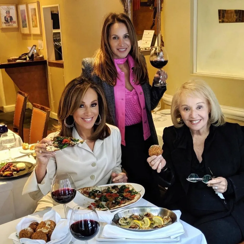 Three women smiling and holding food and wine at a restaurant table.