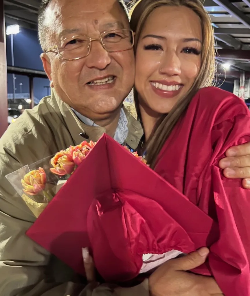 Shukur Aikebaer embraces his daughter, who is wearing a graduation gown and holding flowers and a graduation cap.