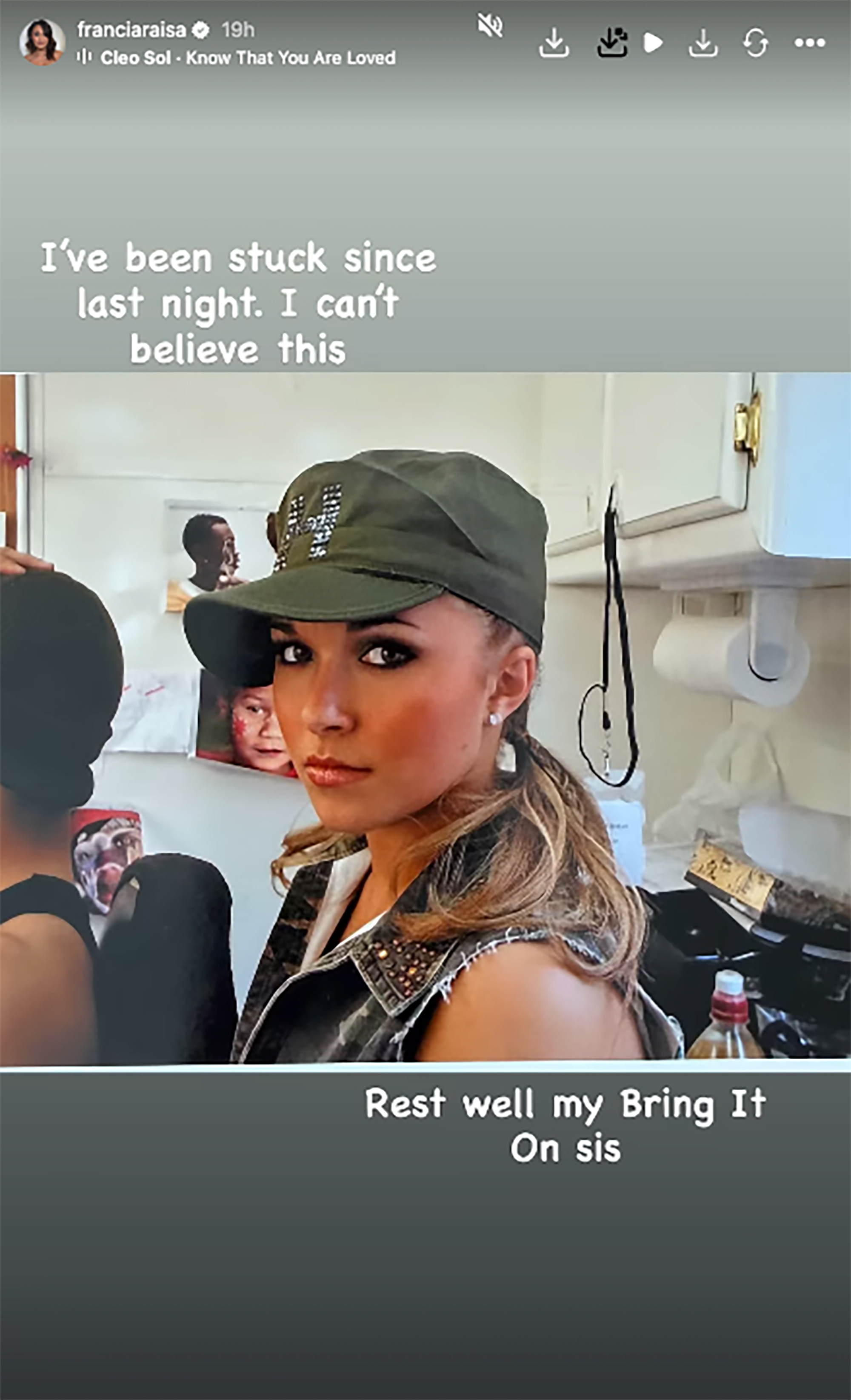 A young woman with a long ponytail, wearing a green cap and a studded vest, looks towards the viewer.