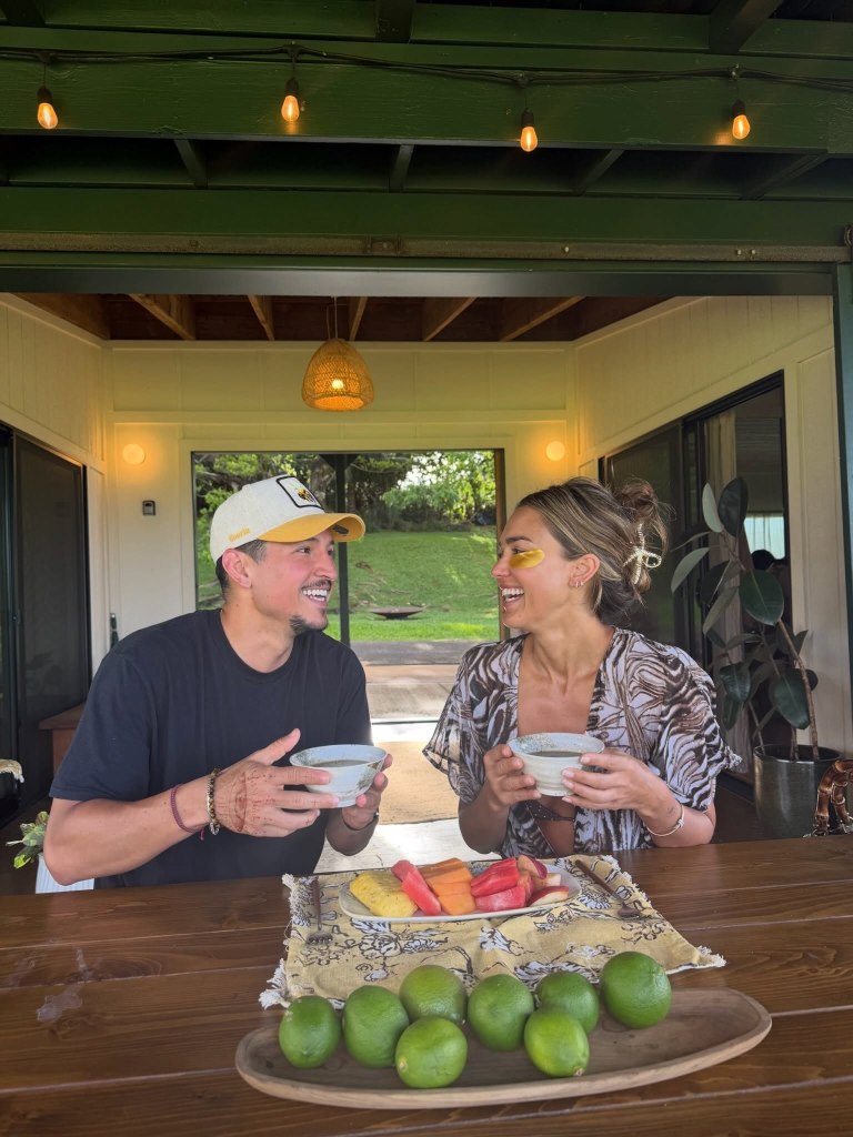 Jessica Alba and a man smiling at each other while holding bowls at a wooden table with fruit and limes.