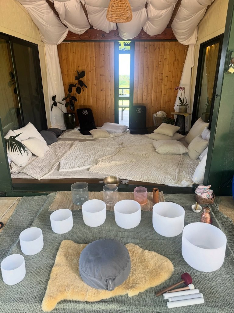 A serene outdoor space set up for meditation with white pillows and blankets, crystal singing bowls, and a yoga cushion.