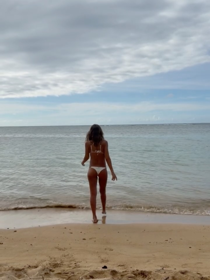 Jessica Alba in a tiny white bikini walking into the ocean from a sandy beach.